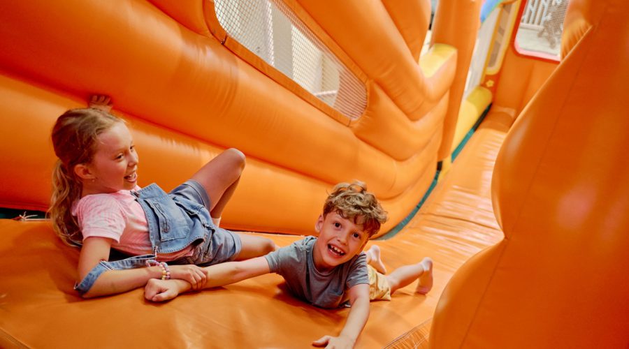 Cute little girl holding her friend by hand while both lying on top of steep orange leather slide and trying not to roll down